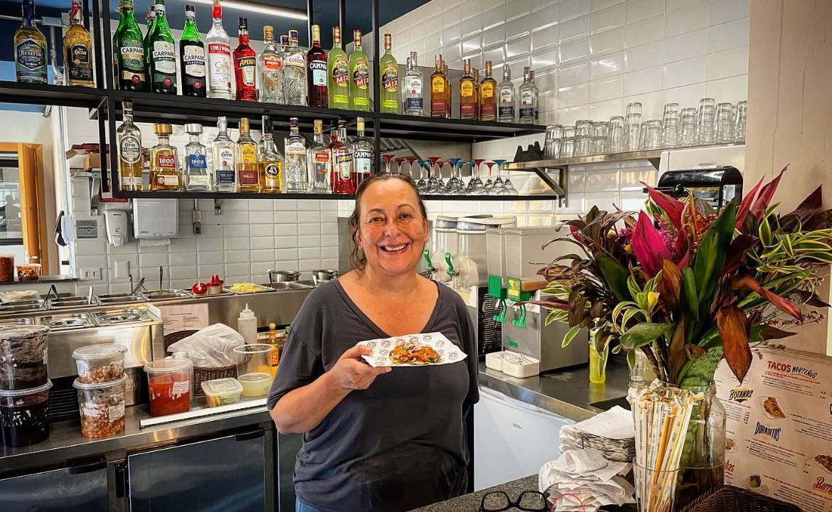 Mulher dentro de cozinha de restaurante com bar atrás segurando um prato de papel com taco.