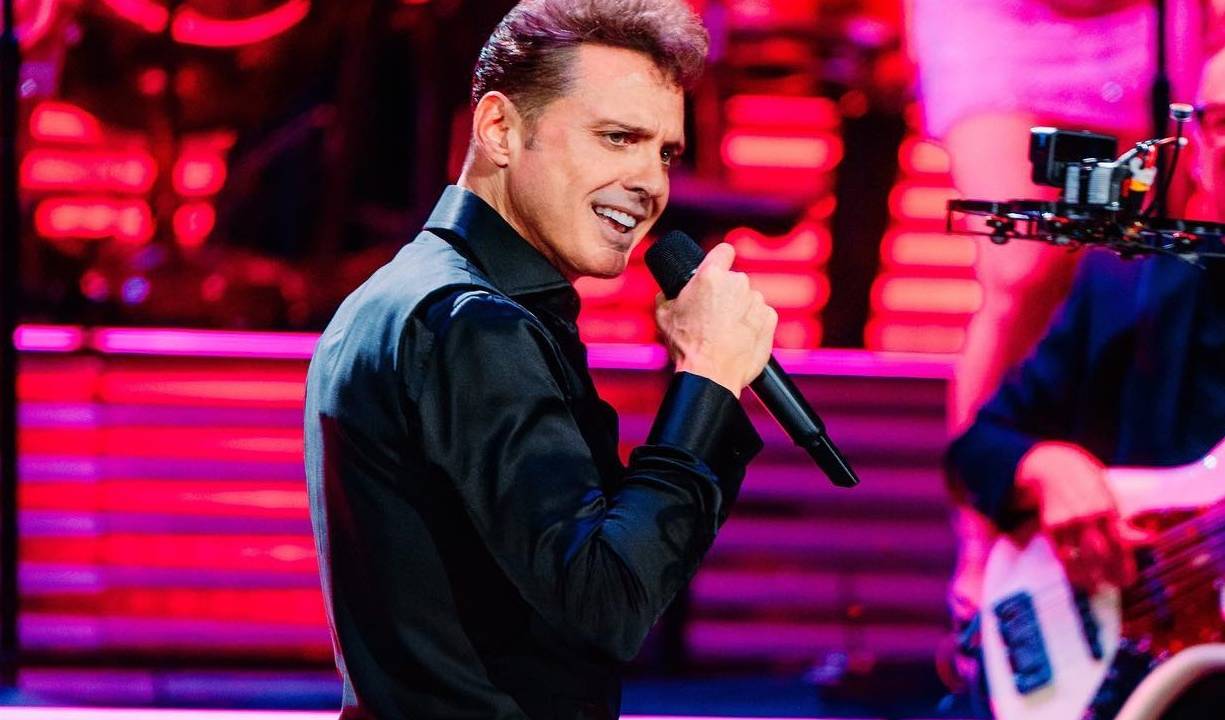 Luis Miguel, homem branco com cabelo castanho em terno de perfil com microfone na mão em palco com holofotes