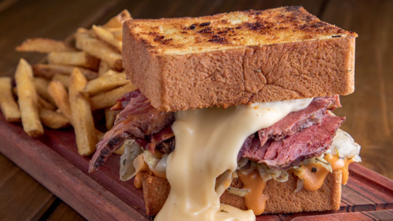 A imagem mostra um sanduíche feito com duas fatias de pão de forma, carne e queijo derretido. Ao fundo, batatas fritas.