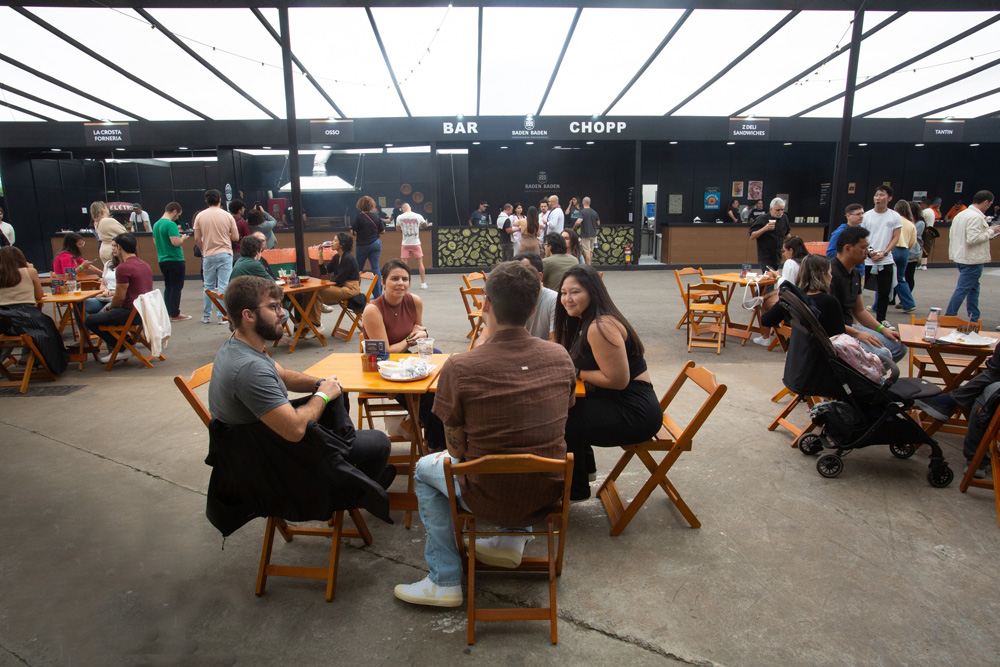 FEIRA DOS CAMPEÕES COMER BEBER Área externa com várias mesas e cadeiras de madeira, pessoas estão sentadas conversando. Ao fundo é possível ver um estande preto.