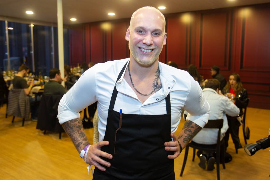 FEIRA DOS CAMPEÕES COMER BEBER 2023 Homem branco sorri para a foto com as mãos na cintura. Ele usa uma camisa branca e um avental preto.