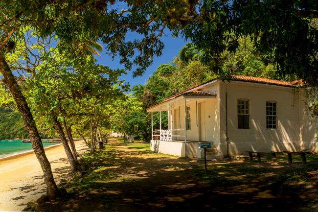 Imagem mostra pequena casa na costa de praia em local arborizado, com grama do lado da casa e areia próxima ao mar