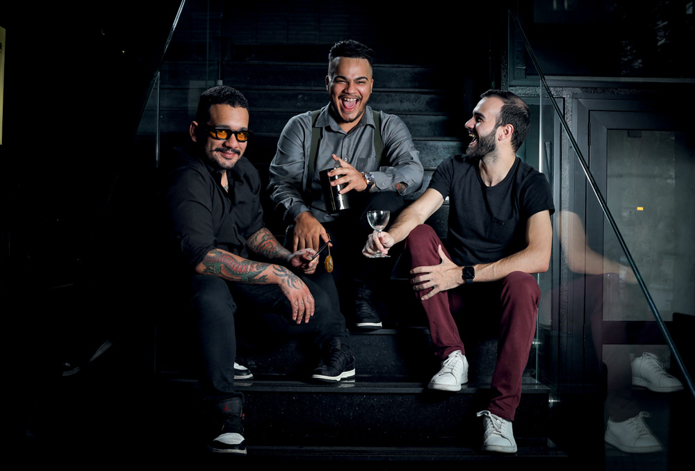 Retrato de três homens sorrindo para a foto. Eles estão sentados em uma escada preta.