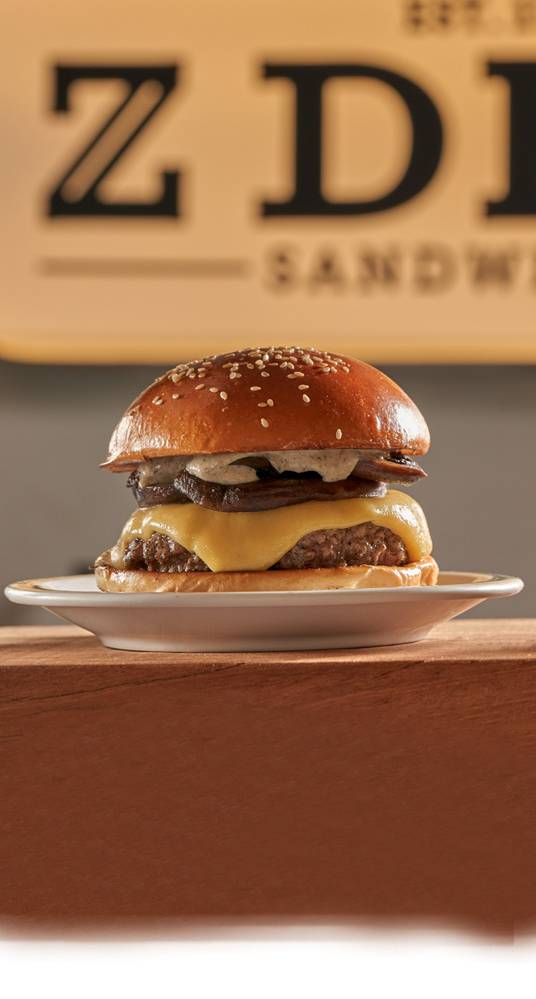 Feira dos Campeões 2023 – Z Deli Sandwiches Hambúrguer com queijo derretido sobre prato de louça branca