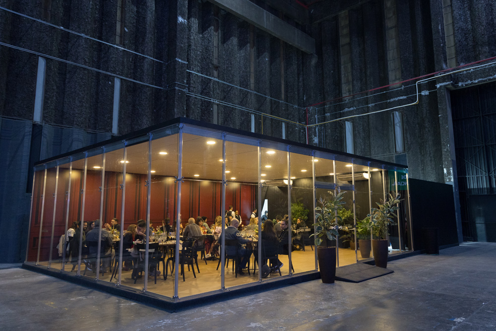 FEIRA DOS CAMPEÕES COMER E BEBER 2023 A imagem mostra uma sala fechada vista por fora com paredes de vidro. Do lado de dentro é possível ver mesas e cadeiras e pessoas sentadas.