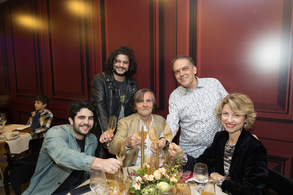 FEIRA DOS CAMPEÕES COMER E BEBER 2023 Cinco pessoas seguram taças de champanhe na mão enquanto posam para a foto e sorriem. Três estão sentadas na frente de uma mesa e duas estão de pé na parte de trás.
