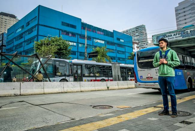 avenida santo amaro ponto de ônibus Imagem mostra jovem em ponto de ônibus olhando para a frente. Dois ônibus passam atrás do rapaz. Ele segura uma mochila