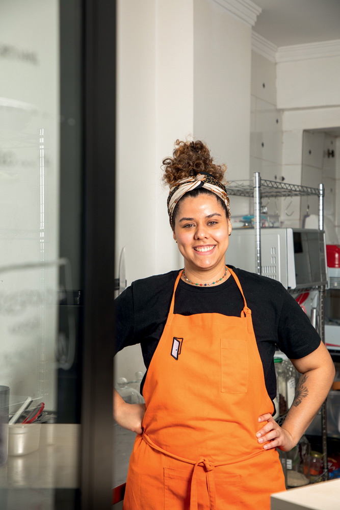 Lauany Carreiro A portinha Retrato de Lauany com camiseta preta e avental laranja. Ela sorri para a foto com as mãos na cintura.