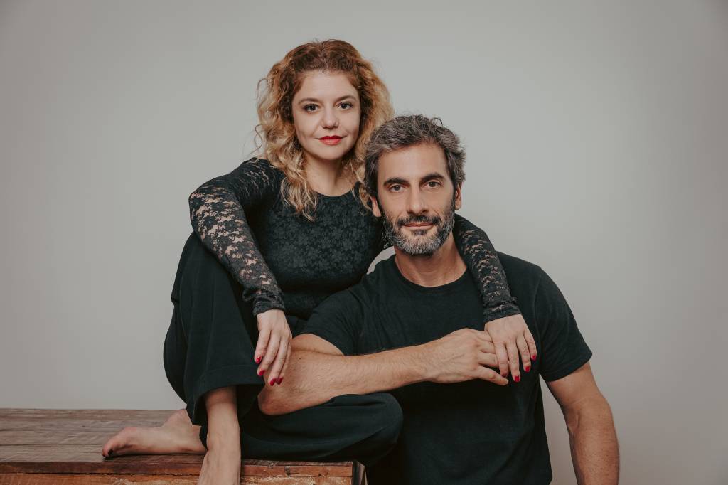 Homem e mulher brancos posam lado a lado, ela mais alta, sentada em cima de uma mesa, com o braço no ombro dele. Ambos vestem peças pretas. Ele tem barba grisalha e ela cabelos loiros.