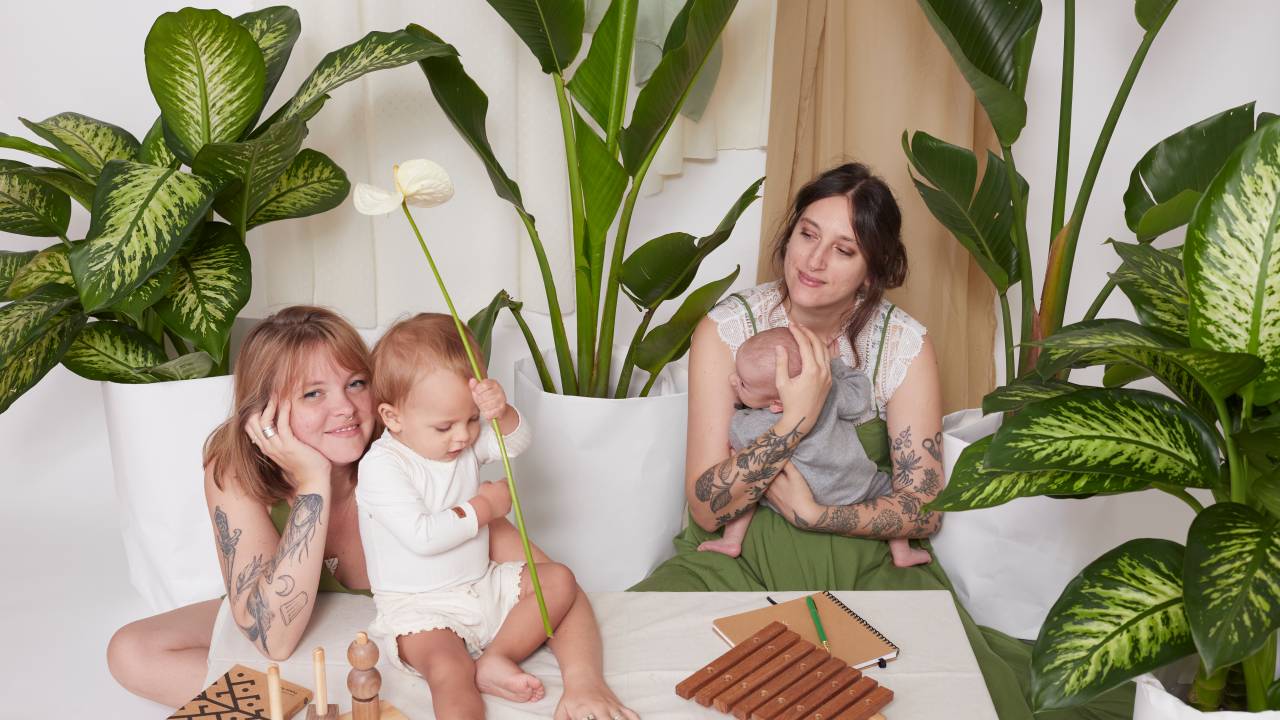 Duas moças posam sentadas no chão com mesa branca. Um bebê está em cima da mesa apoiado à mãe, a mulher loira à esquerda. A da direita segura outro bebê, mais novo, no colo. Elas estão envoltas por plantas da espécie strelitzia em vasos brancos.