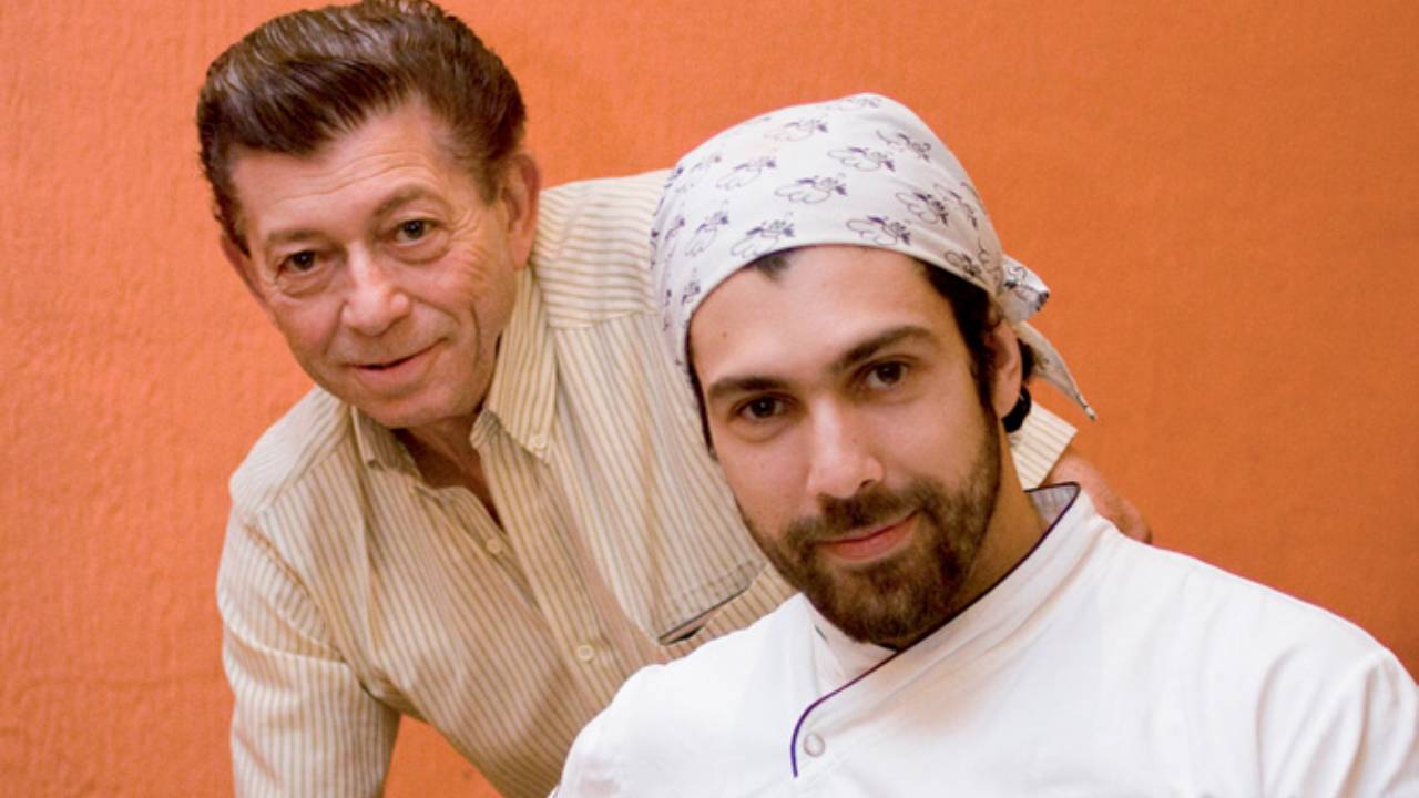 Dois homens, um mais velho atrás e outro de dólmã e lenço na cabeça na frenete.