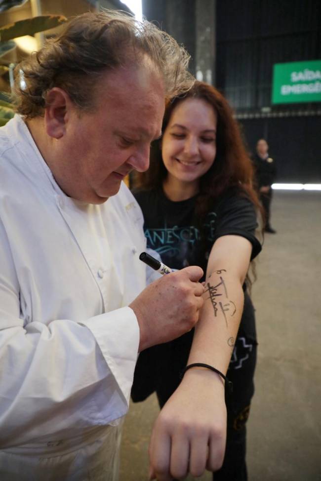 Imagem mostra Jacquin com caneta e jovem de braço estendido. Chef escreve o nome dele no braço da jovem