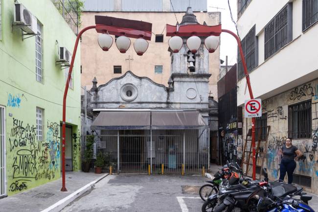 Imagem mostra fim de rua com várias pichações em prédios. Ao fundo, uma pequena igreja, sem cor, com uma torre à direita. Postes são os típicos do bairro da Liberdade