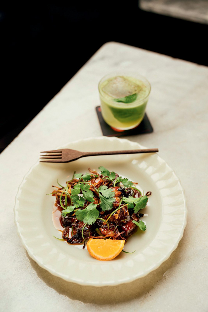 Circuito Comer & Beber – Aconchegante Bar Foto aérea de prato de louça branca com carne glaceada coberta de folhas frescas de coentro com garfo de madeira em cima. Copo baixo com drinque com folha verde em cima.