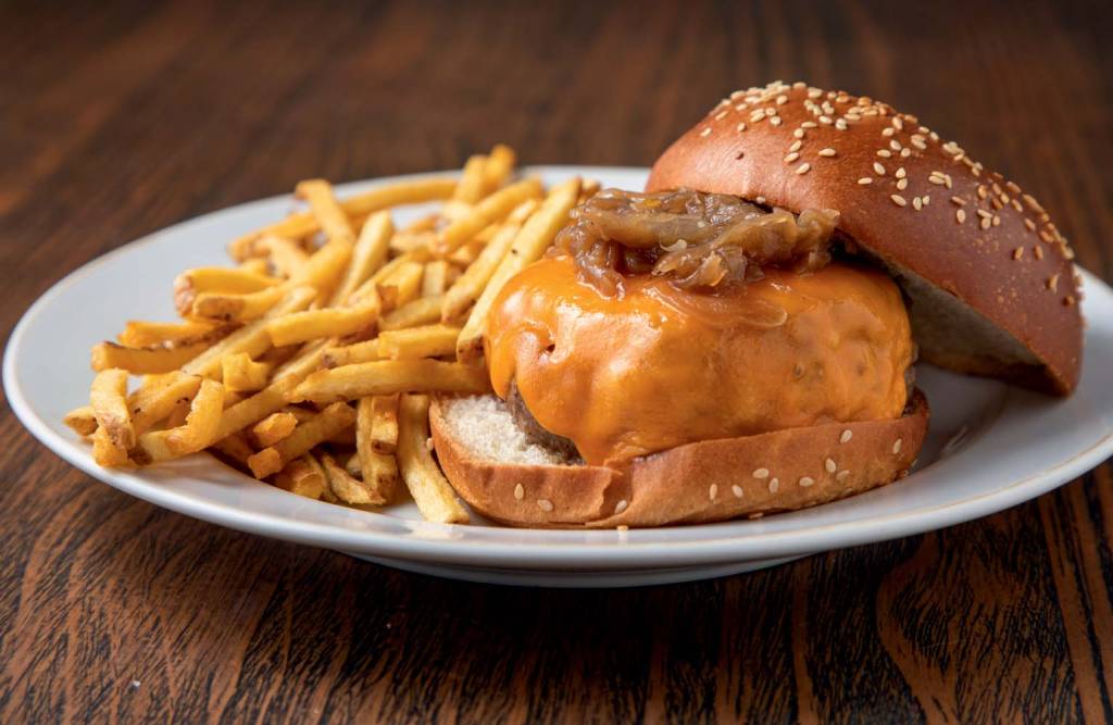 minetta`s burger com fritas _ deBetti Ibirapuera _ restaurantes Sobre uma mesa de madeira há um prato branco com batatas fritas à esquerda e um hambúrguer com queijo cheddar e pão com gergelim do lado.