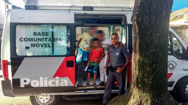 polícia militar Imagem mostra Ronie sorrindo, vestido de farda de policial. Ao lado dele, três crianças posam para foto em cima de van da polícia, uma base comunitária móvel