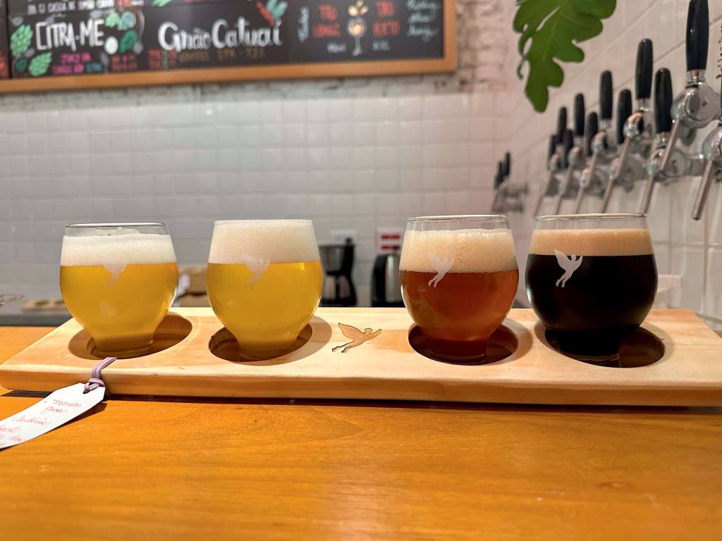Trilha Cervejaria – Itaim Sobre um balcão de madeira, quatro copo com curvatura mais oval apresentam chopes de cores diferentes dentro.