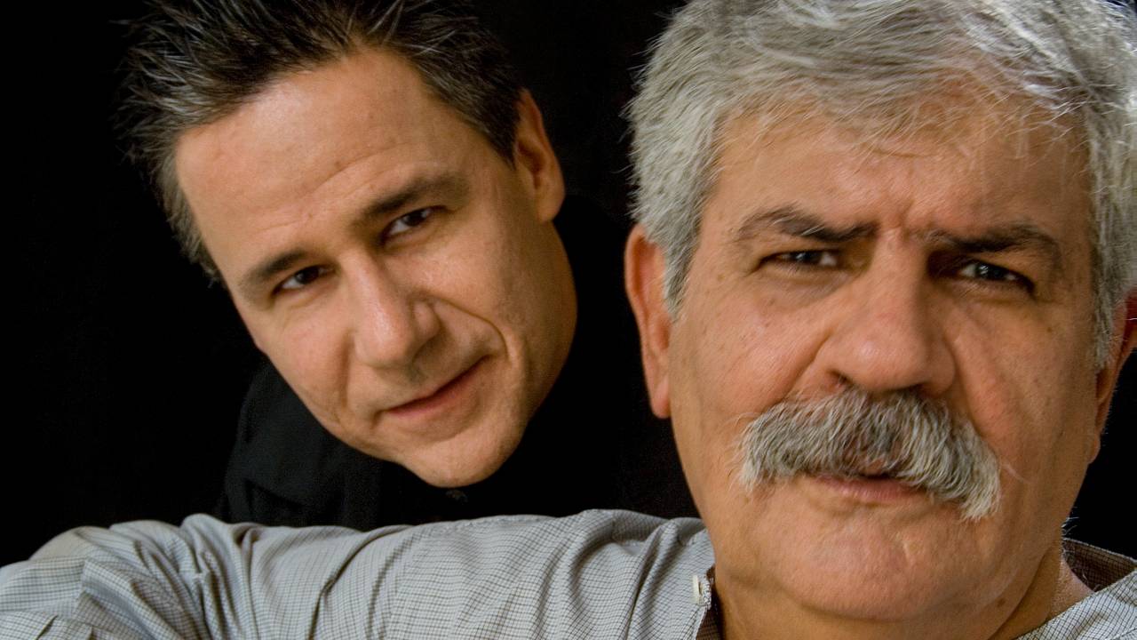 Dois homens brancos posam, um atrás do outro apenas com a cabeça aparecendo, o da frente com violão no colo, grisalho e com bigode.