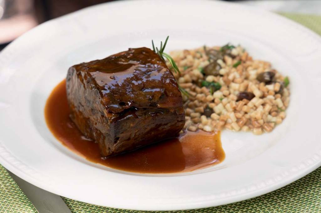 Louça branca com pedaço de carne coberta por molho ao lado de porção de fregola.