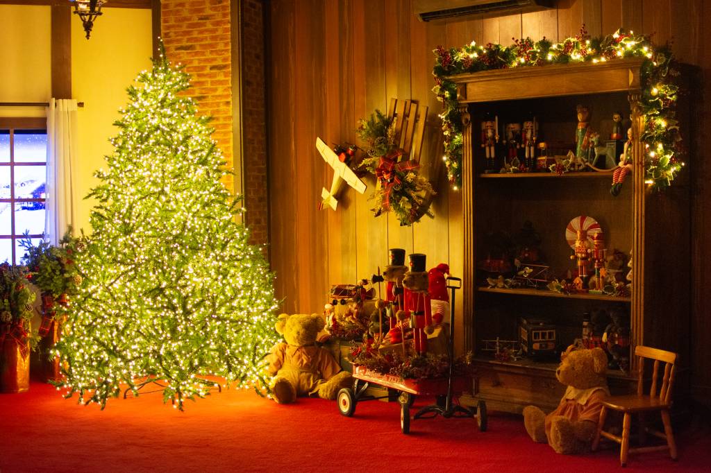 Shopping Higienópolis – Casa do Papai Noel Parte de uma sala de estar com piso vermelho e uma grande árvore de natal com luzes à esquerda. À direita tem uma estante de madeira com várias decorações de natal.