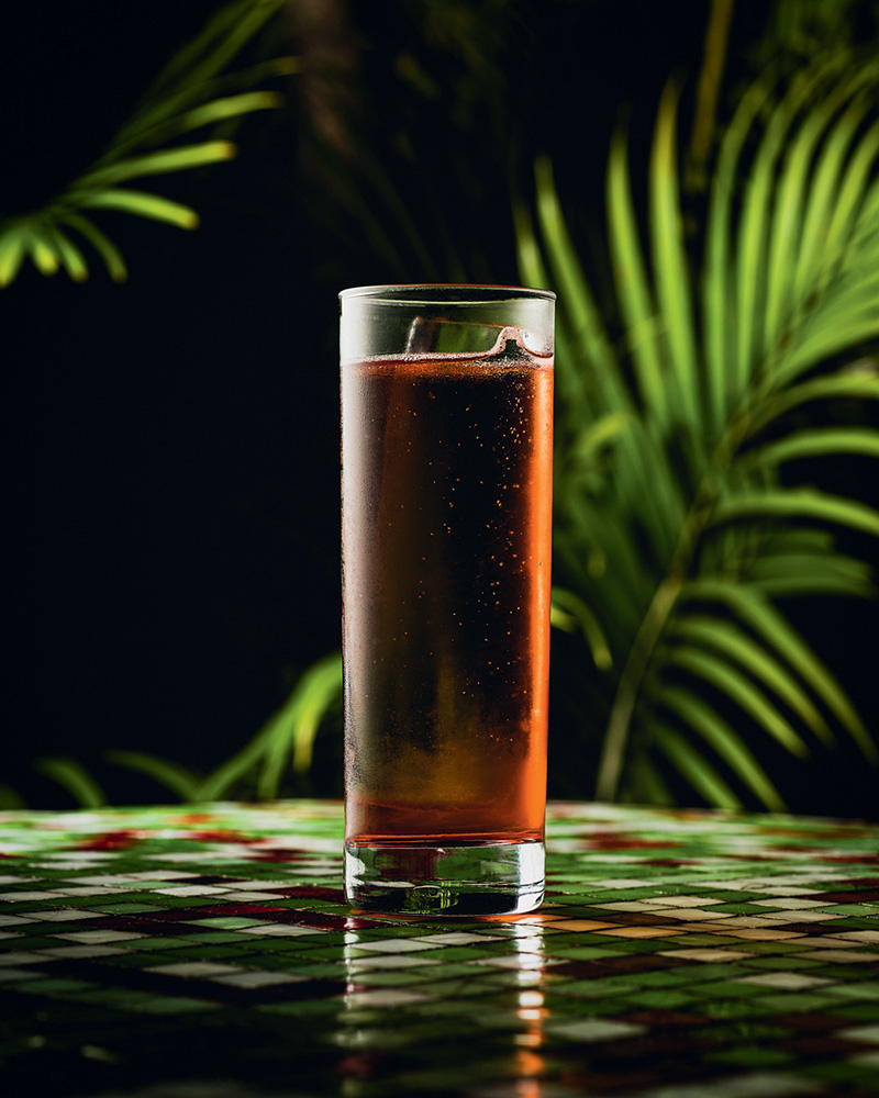 Circuito Comer & Beber – Picco Drinque avermelhado no copo longo com plantas atrás em fundo preto