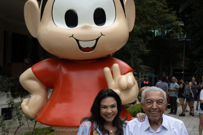 Mauricio de Sousa e sua filha Mônica Sousa posam em pré e sorrindo em frente a uma escultura gigante da Mônica na entrada da Casa das Rosas