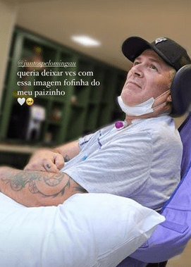 mingau Imagem mostra Mingau sentado, com máscara no queixo. Ele usa boné e está com travesseiro no colo e olha para o canto esquerdo da foto. Ao fundo, uma estante. Texto inserido na foto diz: