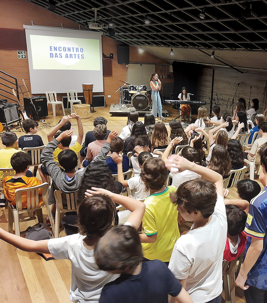 santa cruz Imagem mostra vários alunos olhando palco em que aparece uma menina cantando e outra tocando piano. Ao fundo, pode-se ler no telão