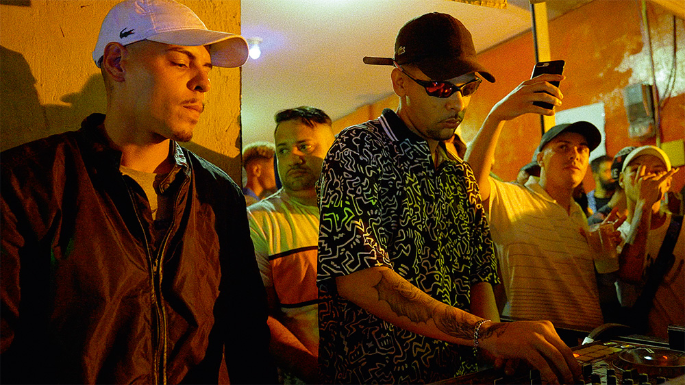 MC Zero K e DJ K Imagem mostra a dupla em um ambiente de festa, com K controlando uma mesa de som enquanto é observado por MC Zero K