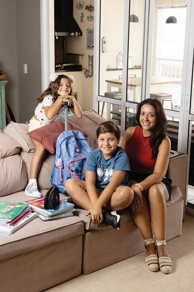 volta às aulas Imagem mostra mãe e duas crianças (uma menina e um menino) em sofá de apartamento, com livros escolares e mochila escolar ao lado. Ao fundo, sacada, com churrasqueira