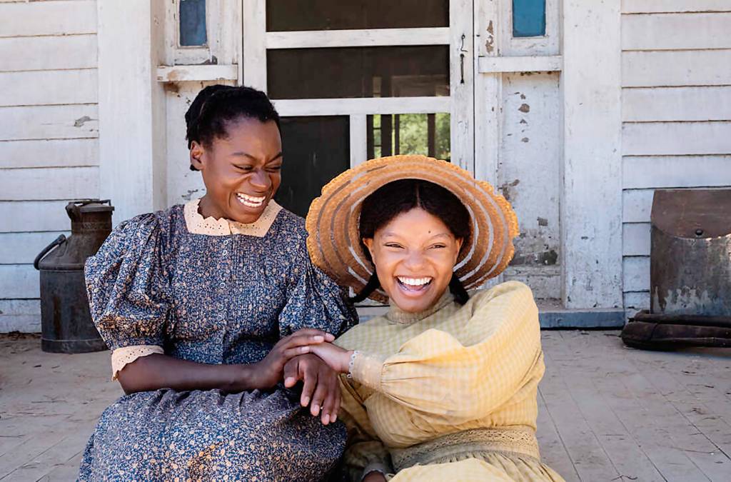 Foto exibe duas mulheres negras com vestidos antigos em escada de terraço de casa com madeira branca. A da direita usa chapéu de palha