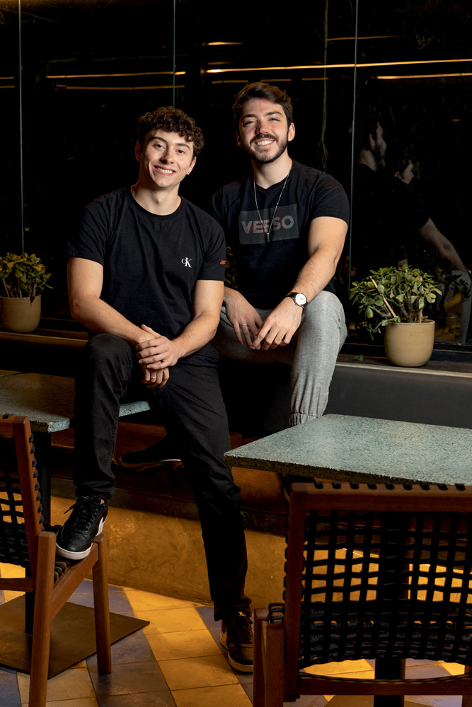 jantar com desconhecidos confra Imagem mostra Guilherme e Lucas sentados em mesa de restaurante, sorrindo. Eles usam camisa preta e calças jeans