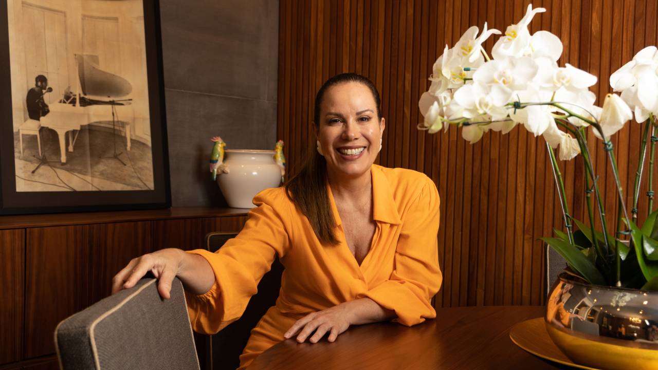 Mulher branca posa sorrindo sentada em mesa com roupa amarela, ao lado de arranjo de orquídeas