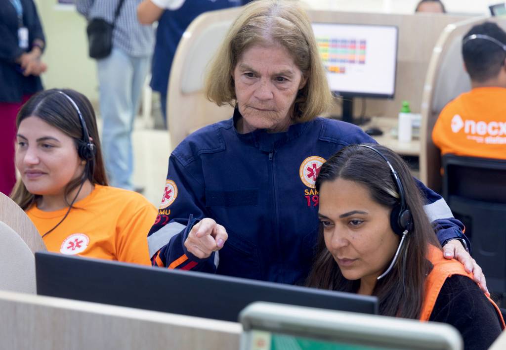 maria jalva samu Imagem mostra Maria Jalva, em pé, apontando para tela de computador de mulher que está sentada na sua direita, com headset para atendimento de chamadas