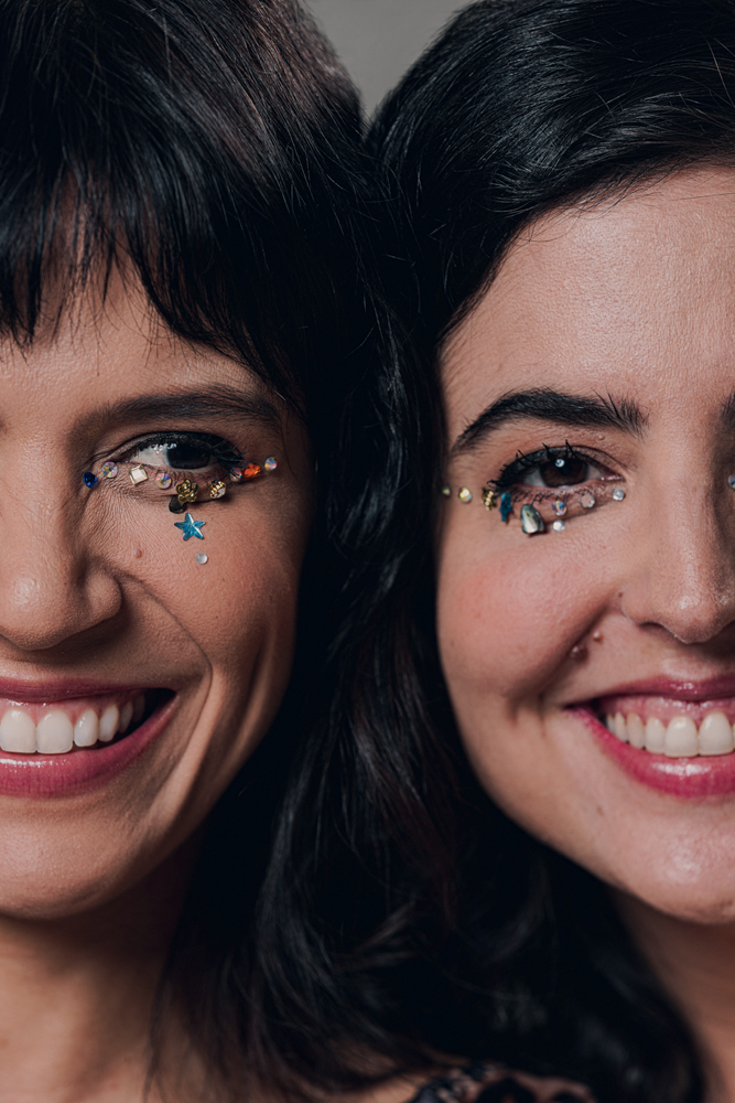 _77A9588_Renata Maciel e Livia La Gatto_Aquela Dupla_Foto Caio Oviedo.jpg Duas mulheres brancas aparecem sorrindo com close só no rosto, ambas com brilhos ao redor de um dos olhos.