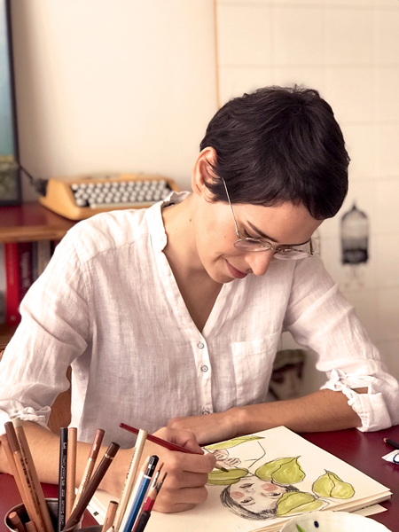 ANNA CUNHA Desenhando Crédito Arquivo pessoal 2.jpg Mulher branca de cabelos curtos posa desenhando em mesa. Veste camisa branca.