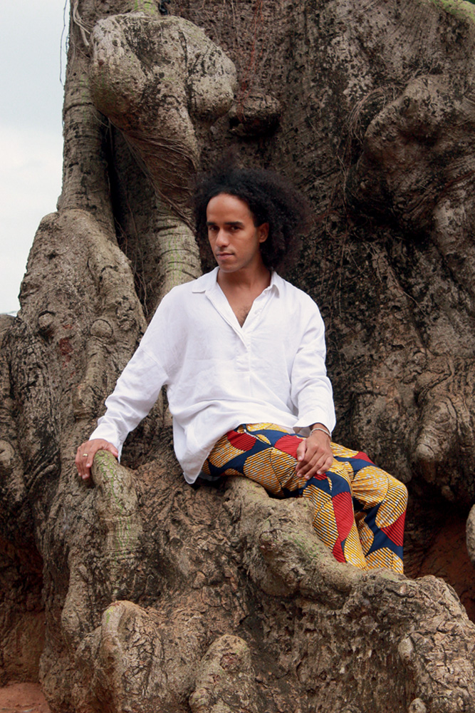Artista Diambe – Crédito Erick Assogba (1).jpg Mulher negra posa sentada em pedra vestindo camisa branca e saia estampada