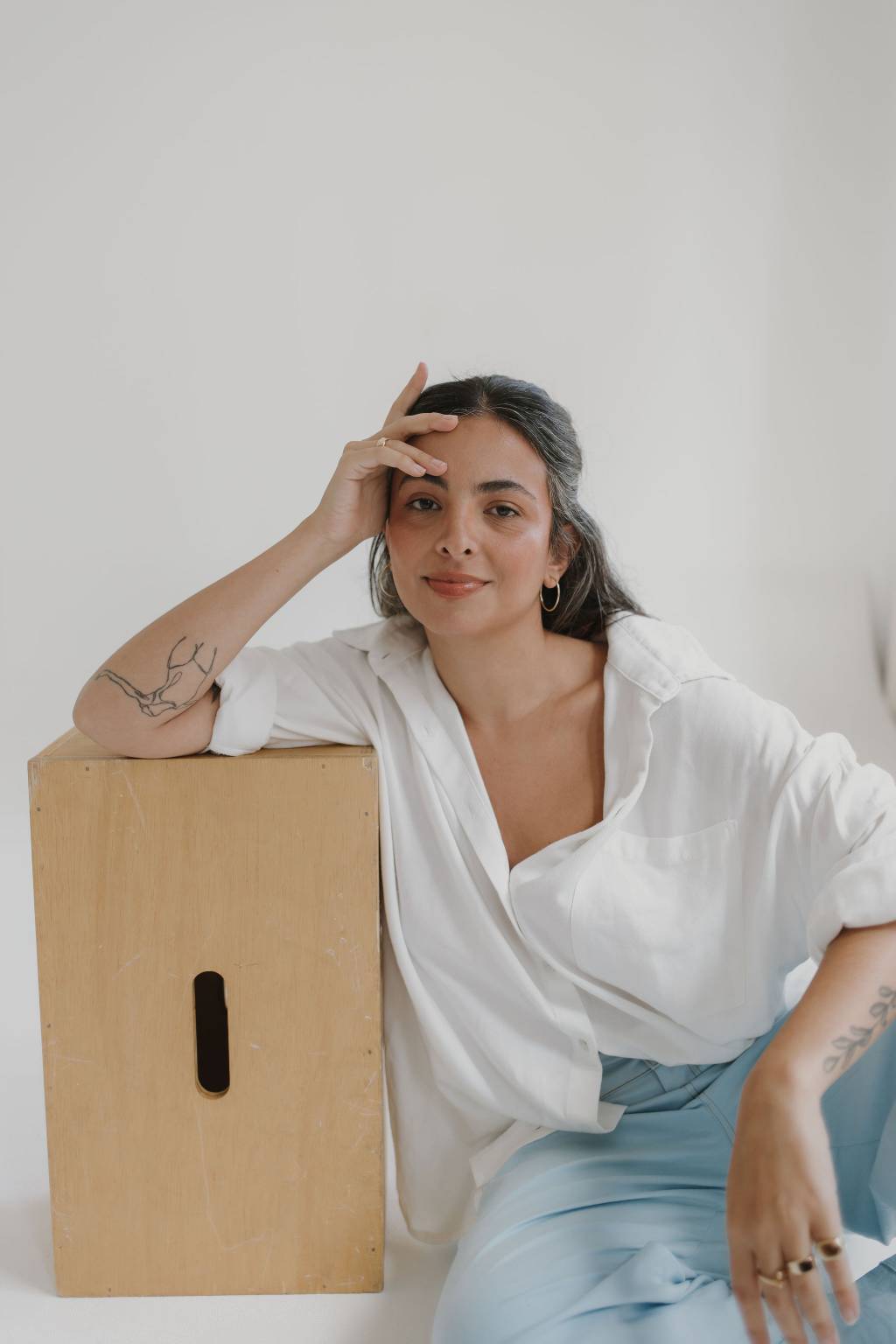 Crédito_ Divulgação Moça branca posa sentada ao lado de caixa de papelão, com um braço apoiado e mão na cabeça. Veste camiseta branca e jeans clara