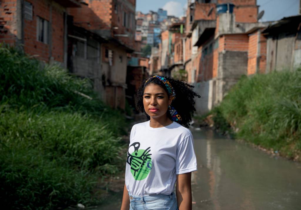 Crédito_ Johnny Miller_Divulgação(2).jpg Mulher negra posa em meio a área com mato e construções com tijolos aparentes. Veste camiseta branca e lenço na cabeça.