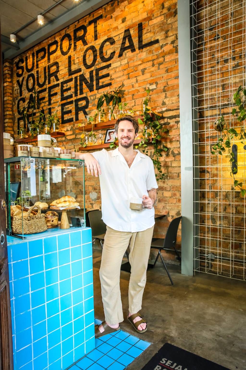 fabian mug.sp Imagem mostra homem em pé em ambiente de tijolinhos ao fundo, com bancada de azulejos azuis ao lado esquerdo da imagem. Ele apoia o braço na bancada, que tem pães e outros produtos, e segura uma xícara de café