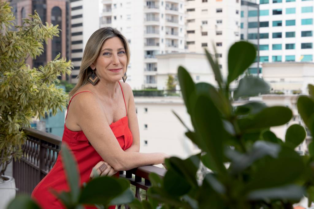 a pesquisadora futurista Sabina Deweik Mulher branca posa em terraço com plantas em primeiro plano e prédios ao fundo. Veste vestido laranja.