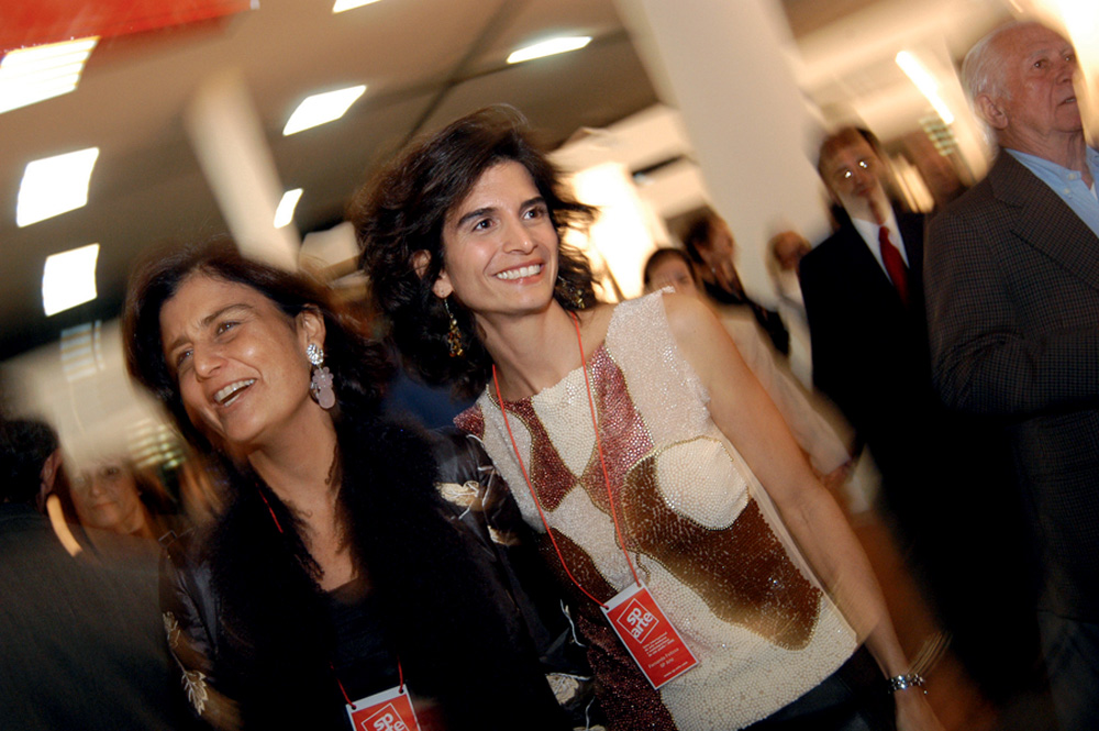 Kátia Avillez e Fernanda Feitosa em 2005 (Divulgação).jpg Duas mulheres brancas sorriem em evento de arte com pilares brancos e pessoas ao fundo. Uma veste regata branca e a outra uma peça preta