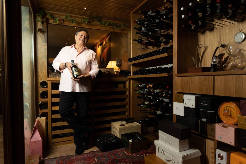 RONNIEVONADEGA13 Senhor branco de cabelos grisalhos sorri segurando garrafa de vinho em adega com prateleiras de madeira e dezenas de garrafas, além de caixas brancas e pretas no chão. Veste camisa bege e calça preta