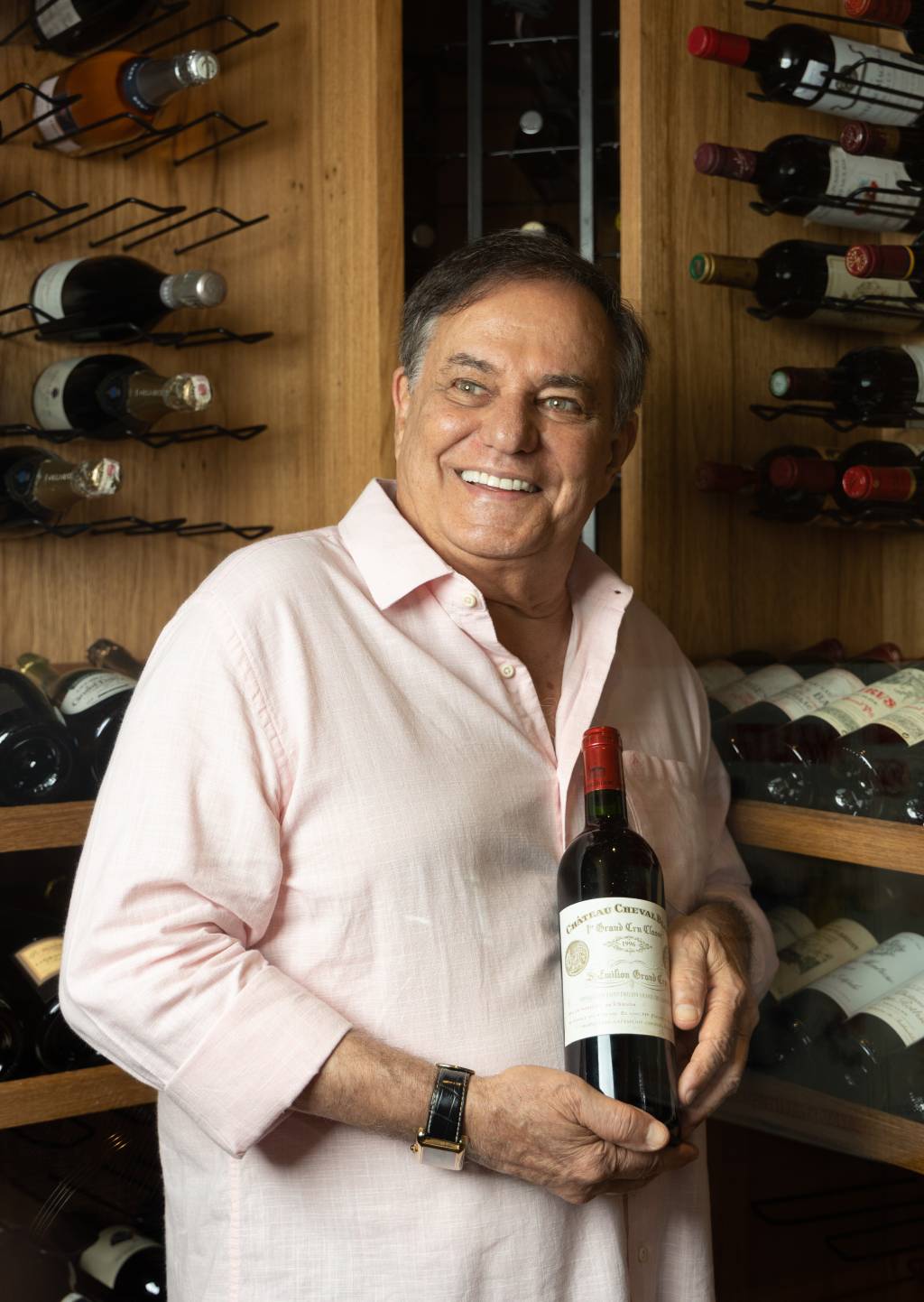RONNIEVONADEGA23 Senhor branco de cabelos grisalhos sorri olhando à esquerda e segurando garrafa de vinho. Veste camisa bege e posa em frente a prateleiras de madeira contendo fileiras de garrafas