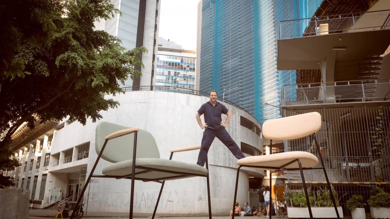 Homem posa com as pernas em duas cadeiras gigantes posicionadas lado a lado.