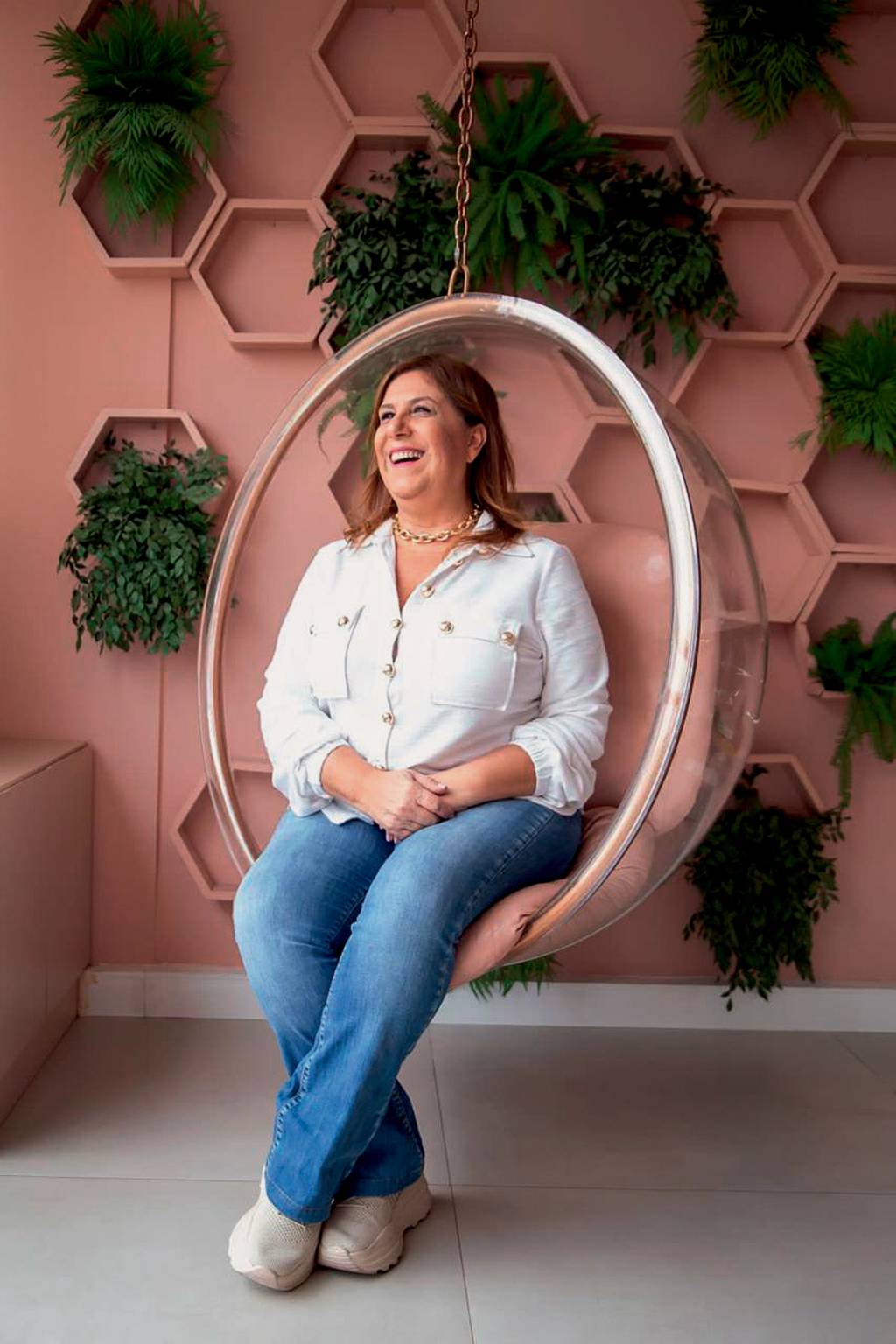 Silvia-Grecco-Crédito-Danutti-de-Carvalho-Divulgação Mulher branca de cabelos lisos posa sentada sorrindo e olhando para a esquerda em cadeira oval transparente. Ao fundo, parede cor de salmão com plantas