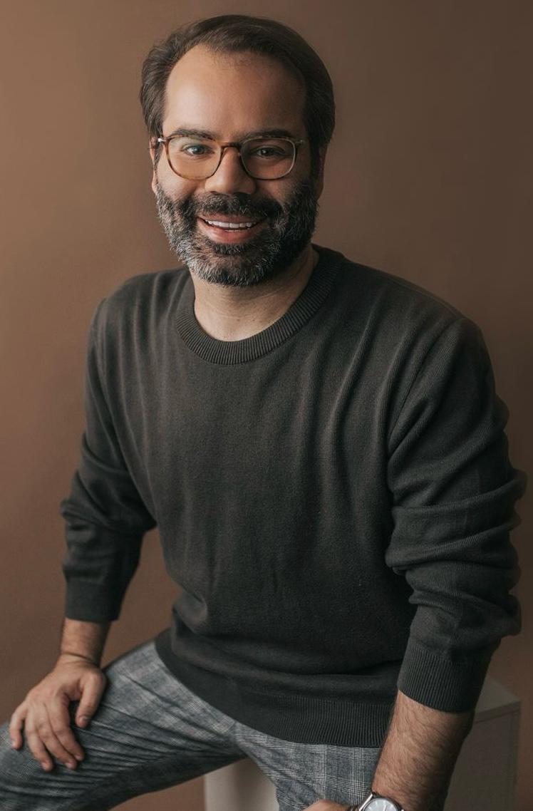 Thiago Braga-Crédito-GSOB-Divulgação (2) Homem de óculos redondo e barba com pelos brancos e pretos posa sorrindo em fundo marrom e vestindo leve suéter preto