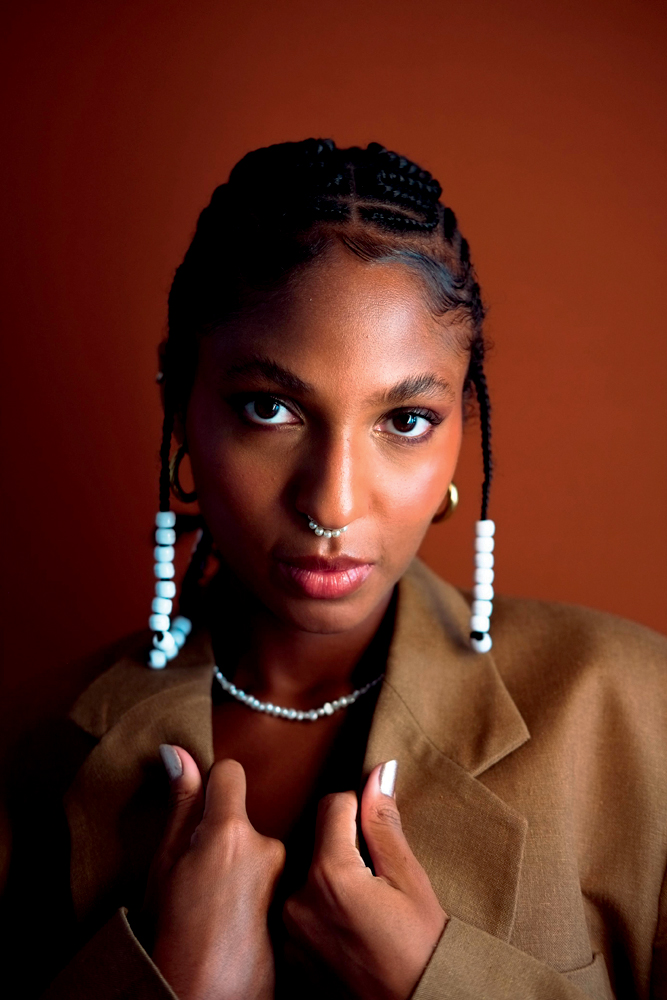 Ayomi Domenica Mulher negra com argolas brancas no cabelo posa encarando a câmera em fundo de tom marrom. Ela veste blazer marrom claro.