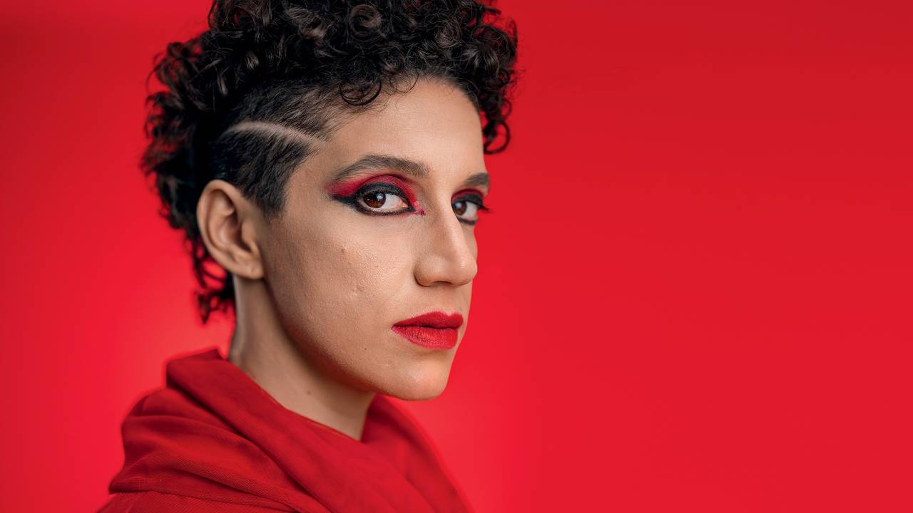 Moça de cabelo preto encaracolado veste peça preta, posa de lado e encara a câmera em fundo vermelho
