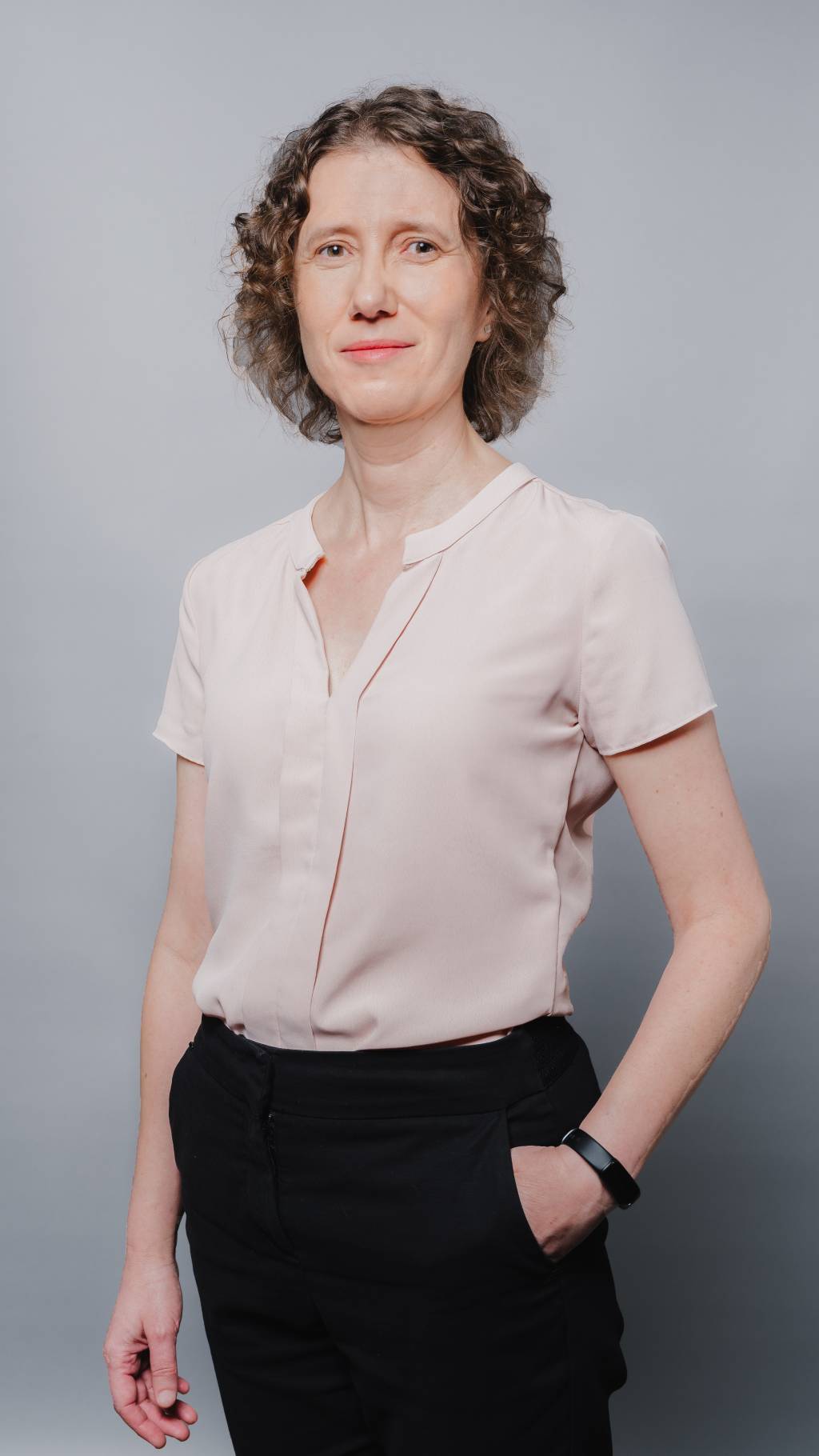 FWIS Mulher cientista de cabelos loiros encaracolados posa com blusa bege e calça preta com uma mão no bolso